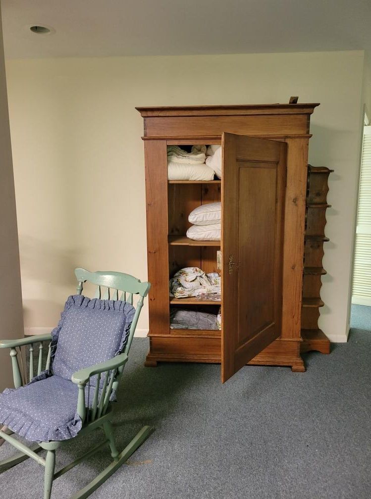 Wooden wardrobe with an open door, stacked folded linens, next to a rocking chair.