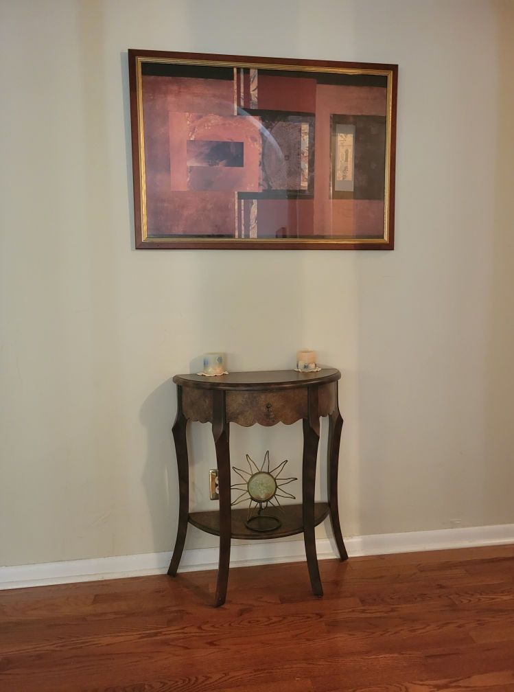 Artwork above a small wooden table against a beige wall and hardwood floor.