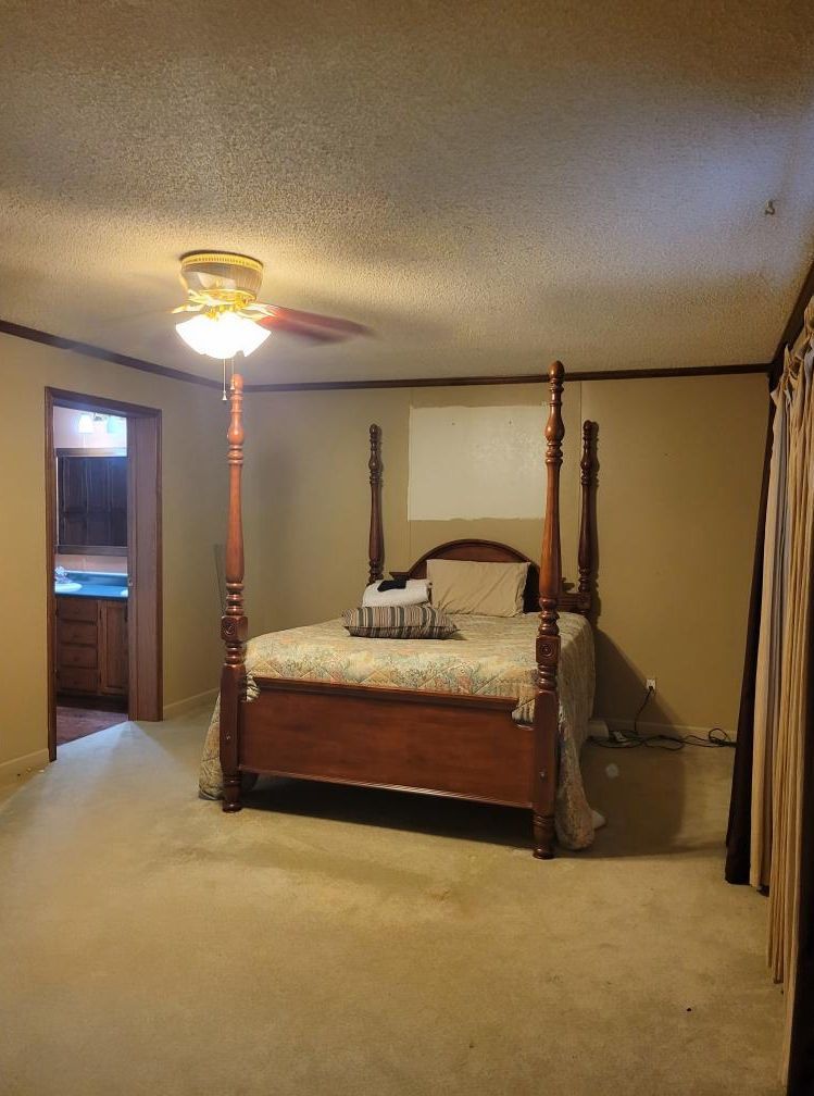 Bedroom with a four-poster bed, ceiling fan, and access to another room. Light carpet and beige walls.