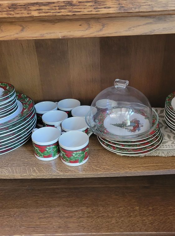 Christmas-themed dishware set in a wooden cabinet. Includes plates, cups, covered serving dish, with red and green accents.