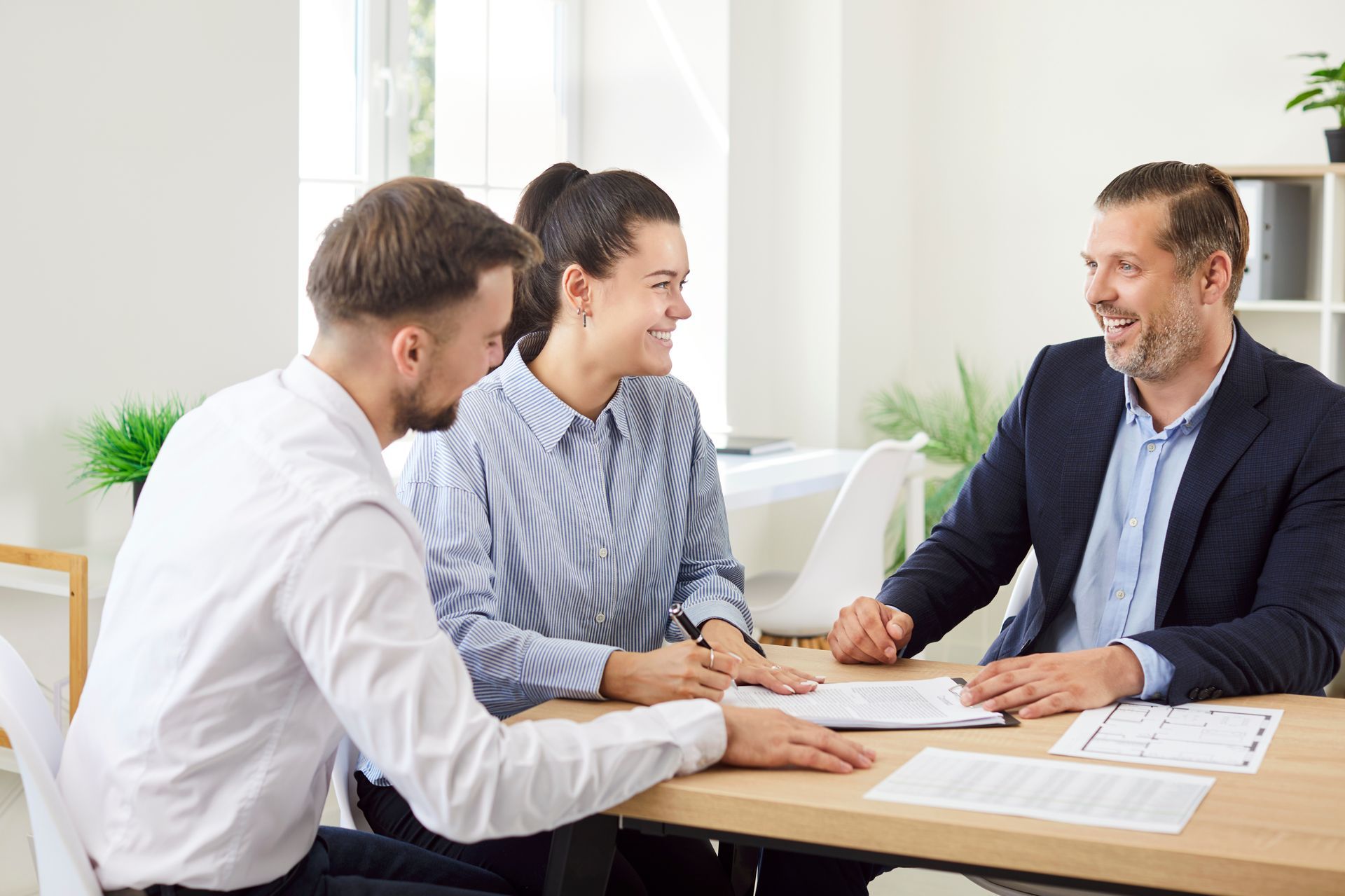 Couple signing documents with a smiling professional at a desk. Light, bright office setting.
