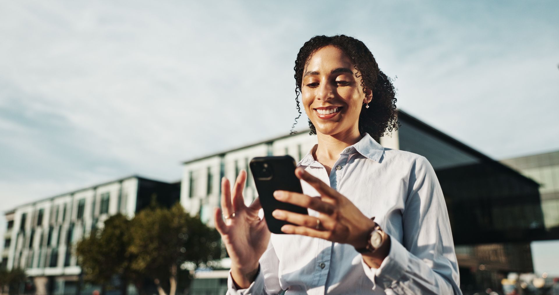 Woman smiling, using a smartphone outdoors, with buildings in the background.