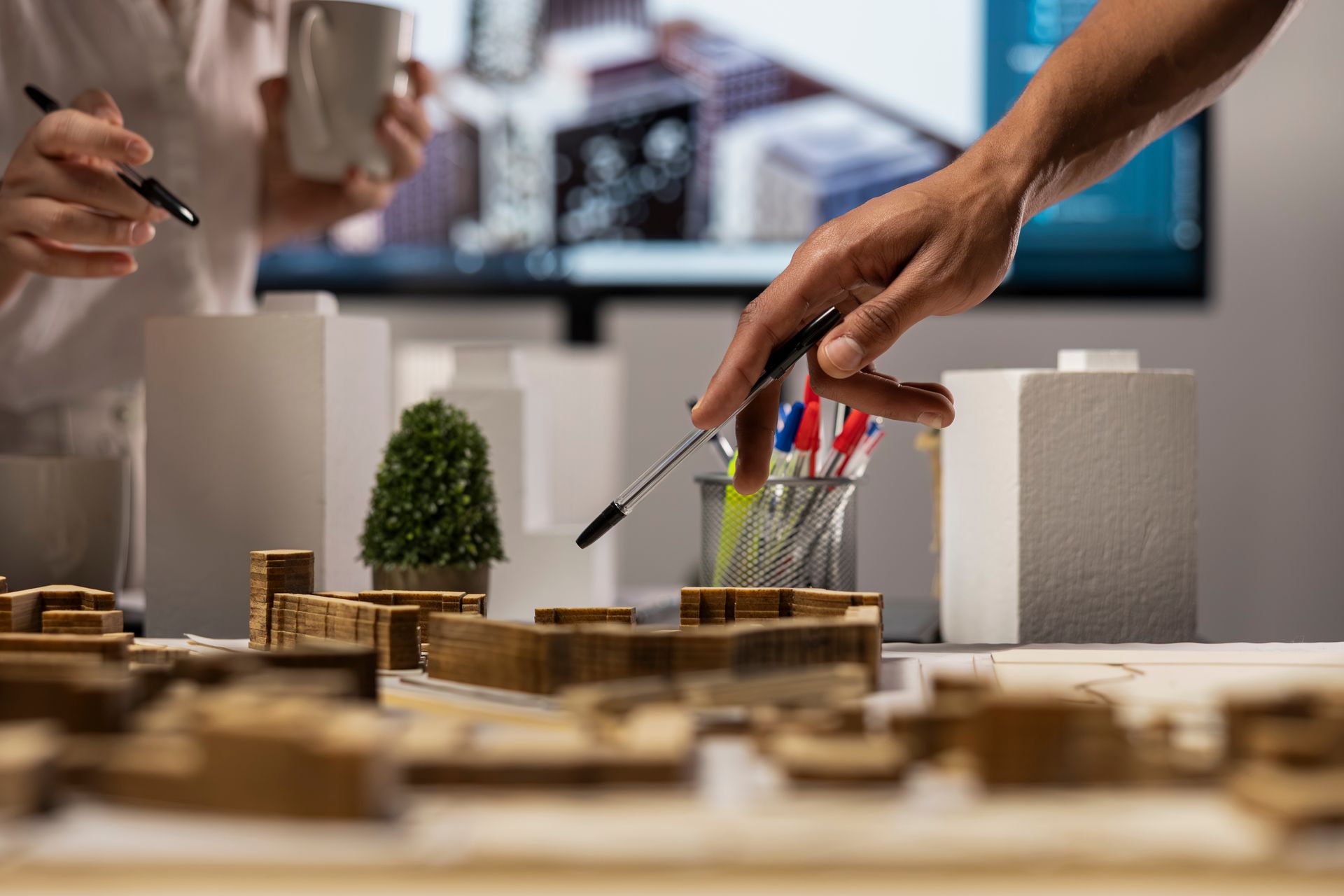 Hands pointing to architectural models; blurry background of a computer screen and person holding a mug.