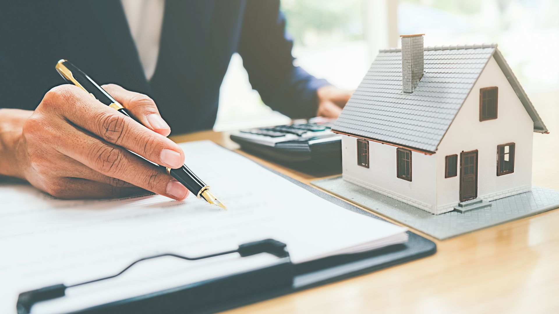 Person signing document with a pen, next to a miniature house and calculator on a desk.