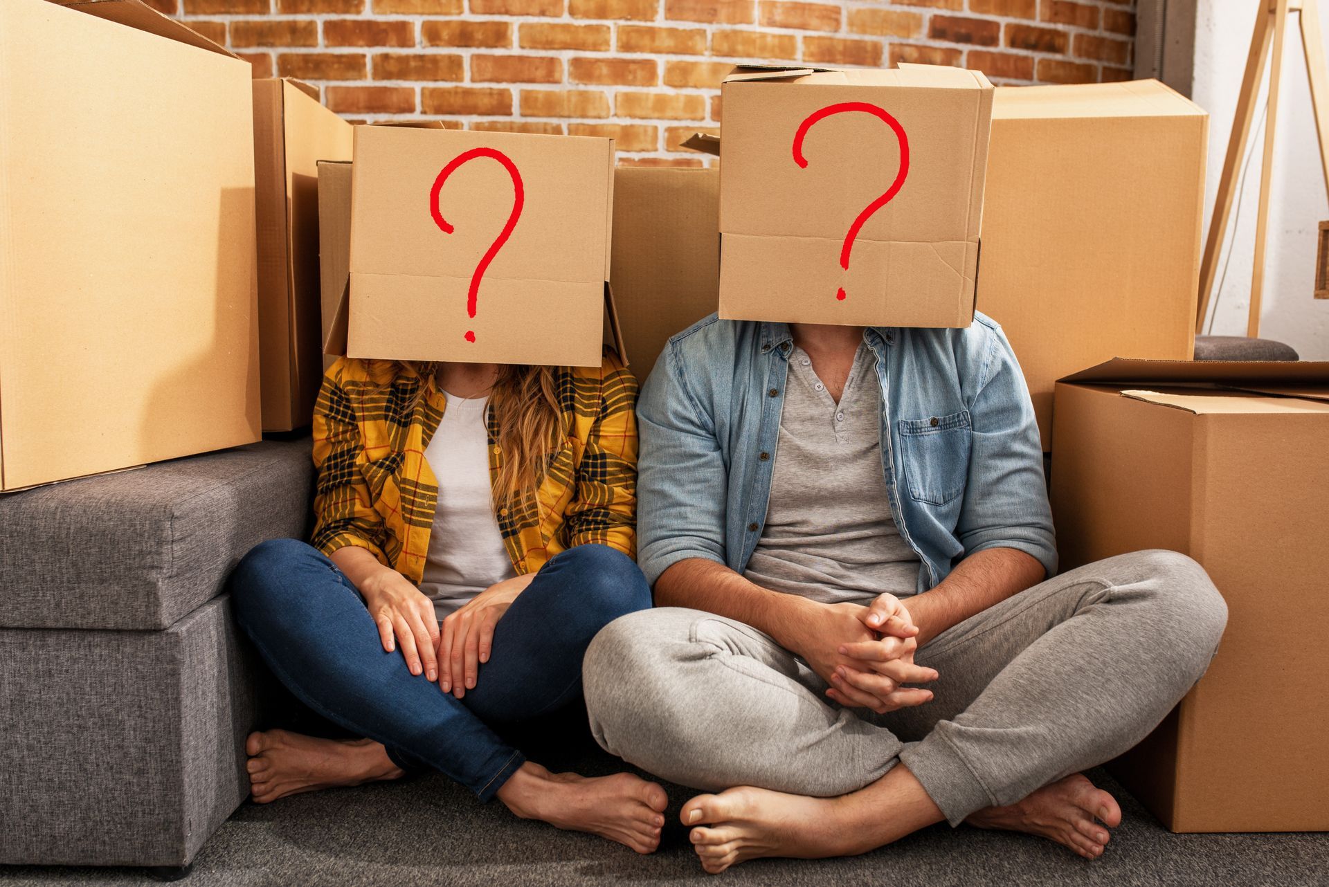 Two people sit with cardboard boxes over their heads, question marks drawn on them, surrounded by moving boxes. Two people sit with cardboard boxes over their heads, question marks drawn on them, surrounded by moving boxes.