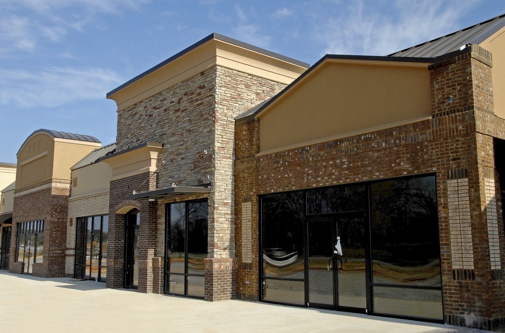 Empty brick and tan-colored storefronts with large glass windows under a clear blue sky.