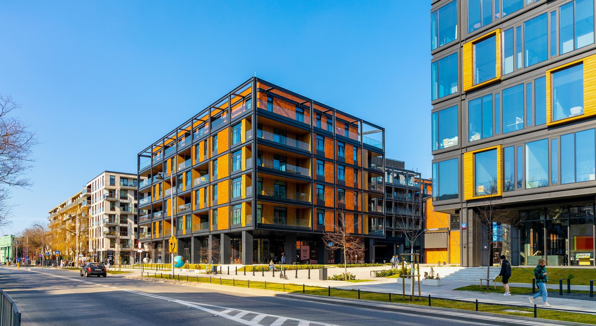 Modern apartment buildings with orange and yellow accents line a city street under a clear blue sky.