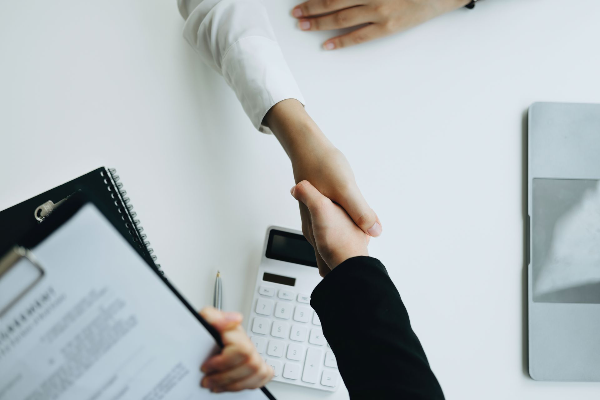 Two people shaking hands over a white table with a clipboard, calculator, and laptop nearby. Two people shaking hands over a white table with a clipboard, calculator, and laptop nearby.