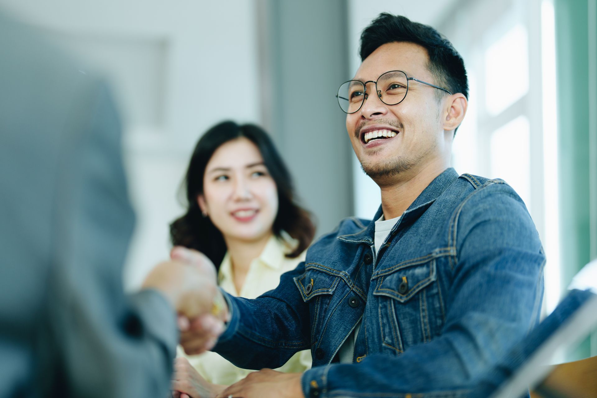 Man in denim jacket shaking hands with another person, smiling. Woman smiles in the background.