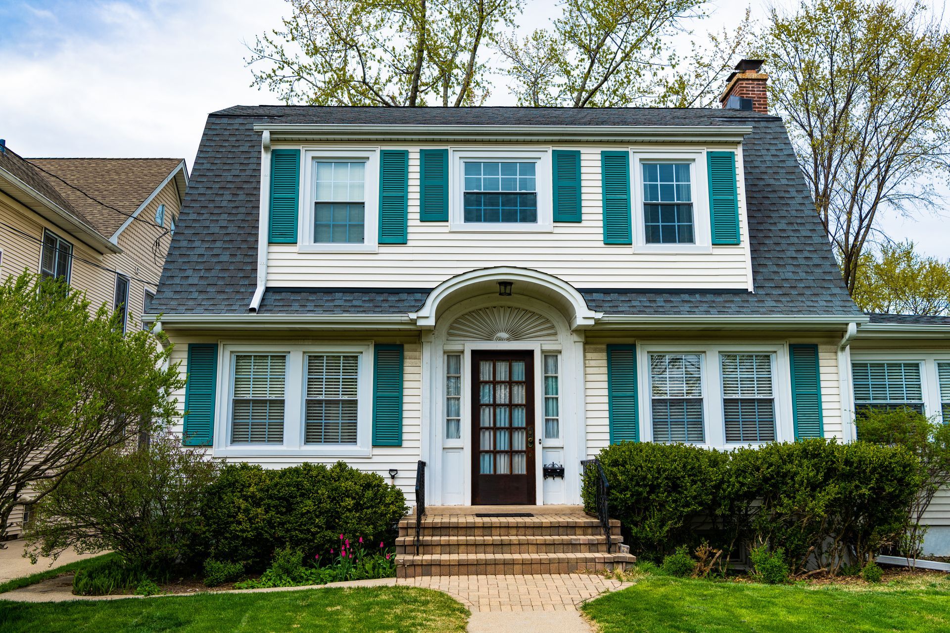 White house with teal shutters, dark roof, brick steps, and bushes.