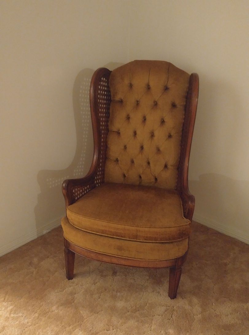 Mustard-colored tufted wingback chair with wooden frame and cane sides, in a corner on a brown carpet.
