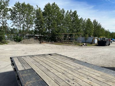 Gravel lot with a wooden trailer in the foreground, trees and structures in the background under a blue sky.