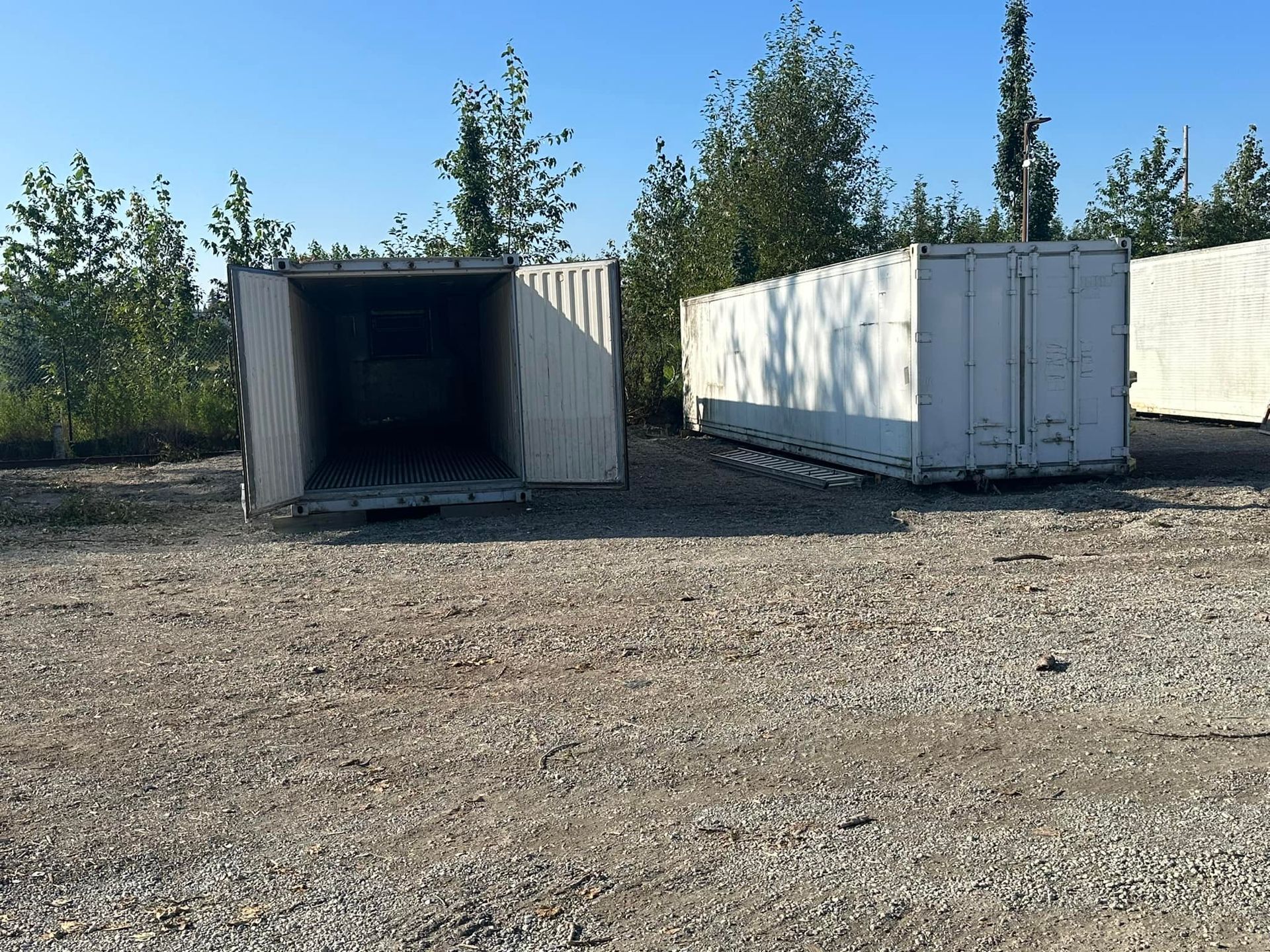 Two white shipping containers on gravel, one with open doors, trees in the background.
