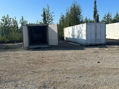 Two white shipping containers on gravel, one with open doors, trees in the background.