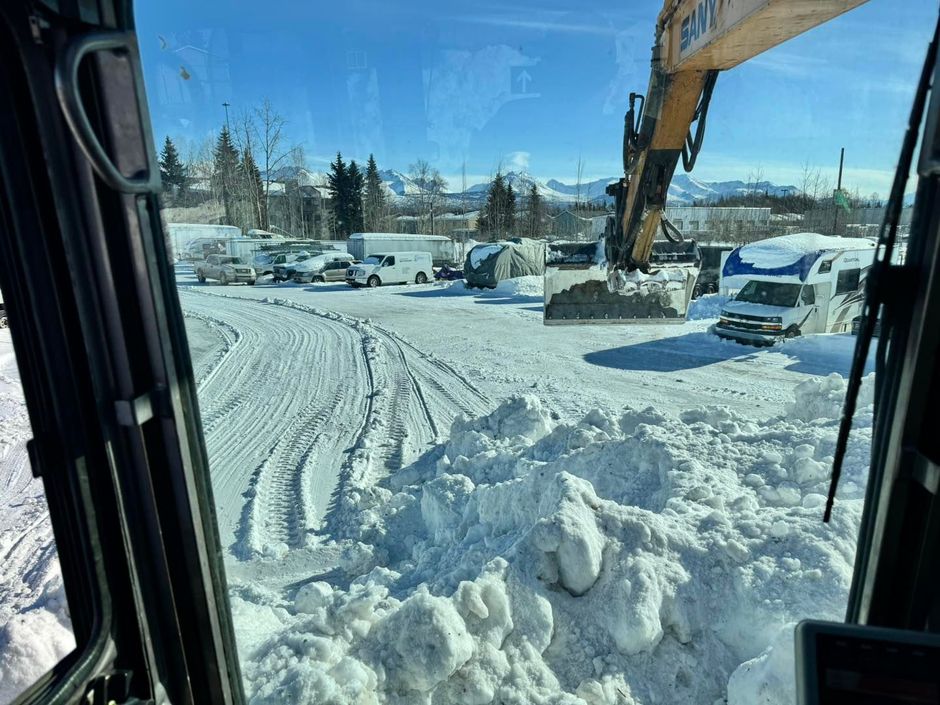 View from excavator cab clearing snow, parked vehicles and mountains in background.