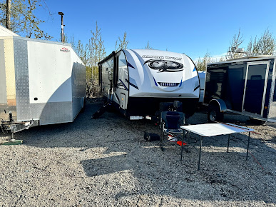 RV parked between two trailers on a gravel lot; table and equipment in front.