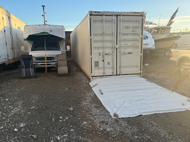 Shipping container with lowered door and white tarp, next to an RV. Outdoors.