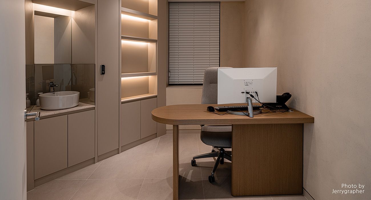 A modern home office with a beige desk, ergonomic chair, built-in shelves with warm lighting, and an integrated sink area.