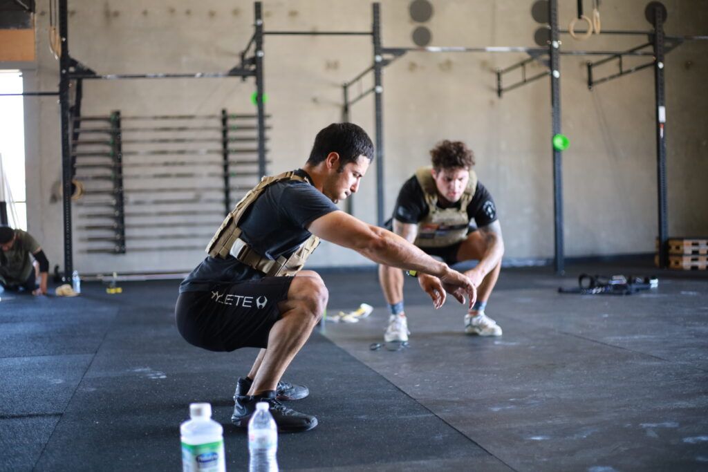 Two men are squatting on the floor in a gym.