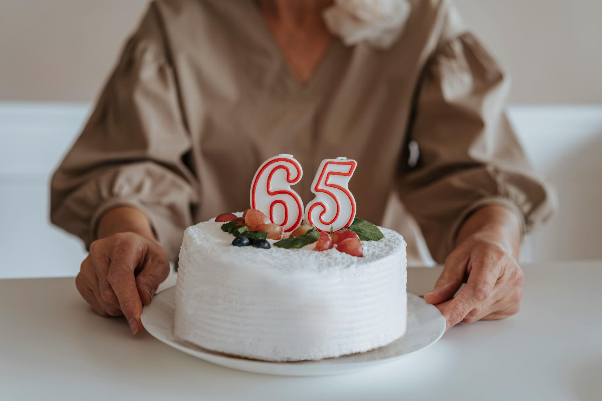 An elderly woman is holding a birthday cake with candles in the shape of the number 65.