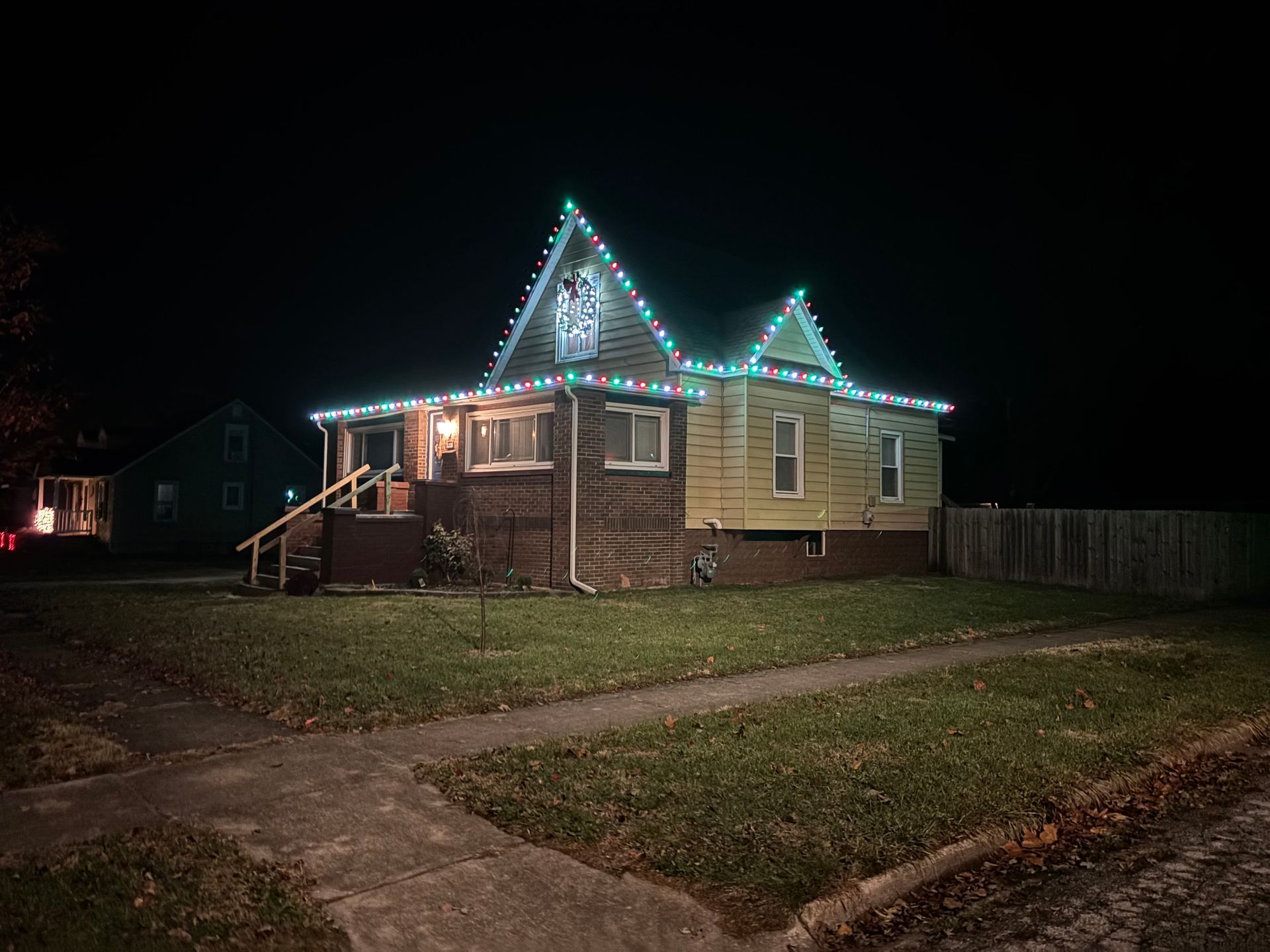 House with green, red and cool white Christmas lights on roof, wreath decoration in center, at night.