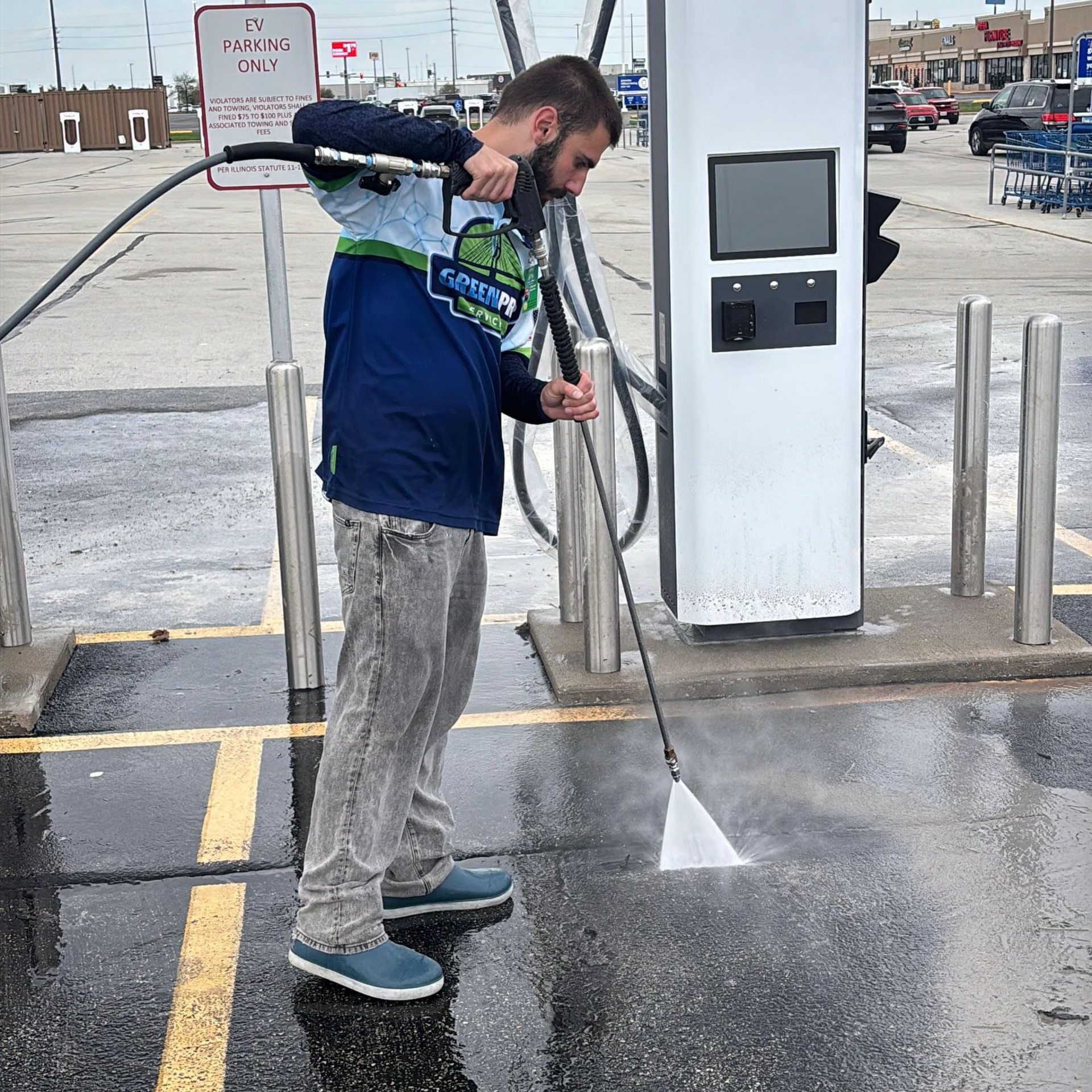 Man using a pressure washer at an EV charging station, spraying water on the pavement.