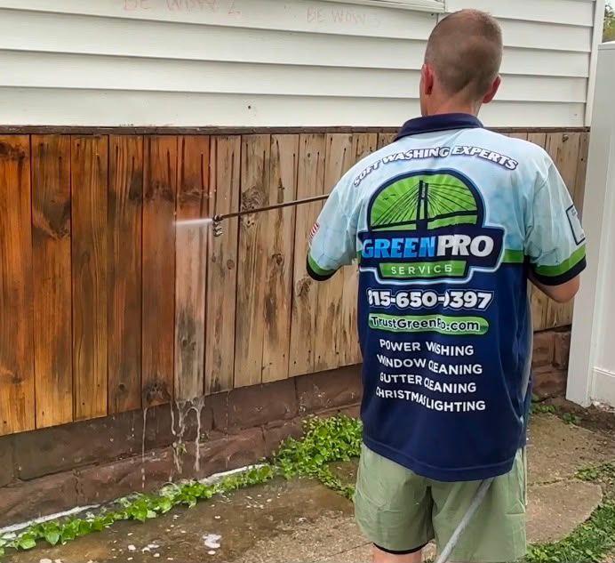 Worker in a Green Pro uniform pressure washing a wood-sided house exterior.