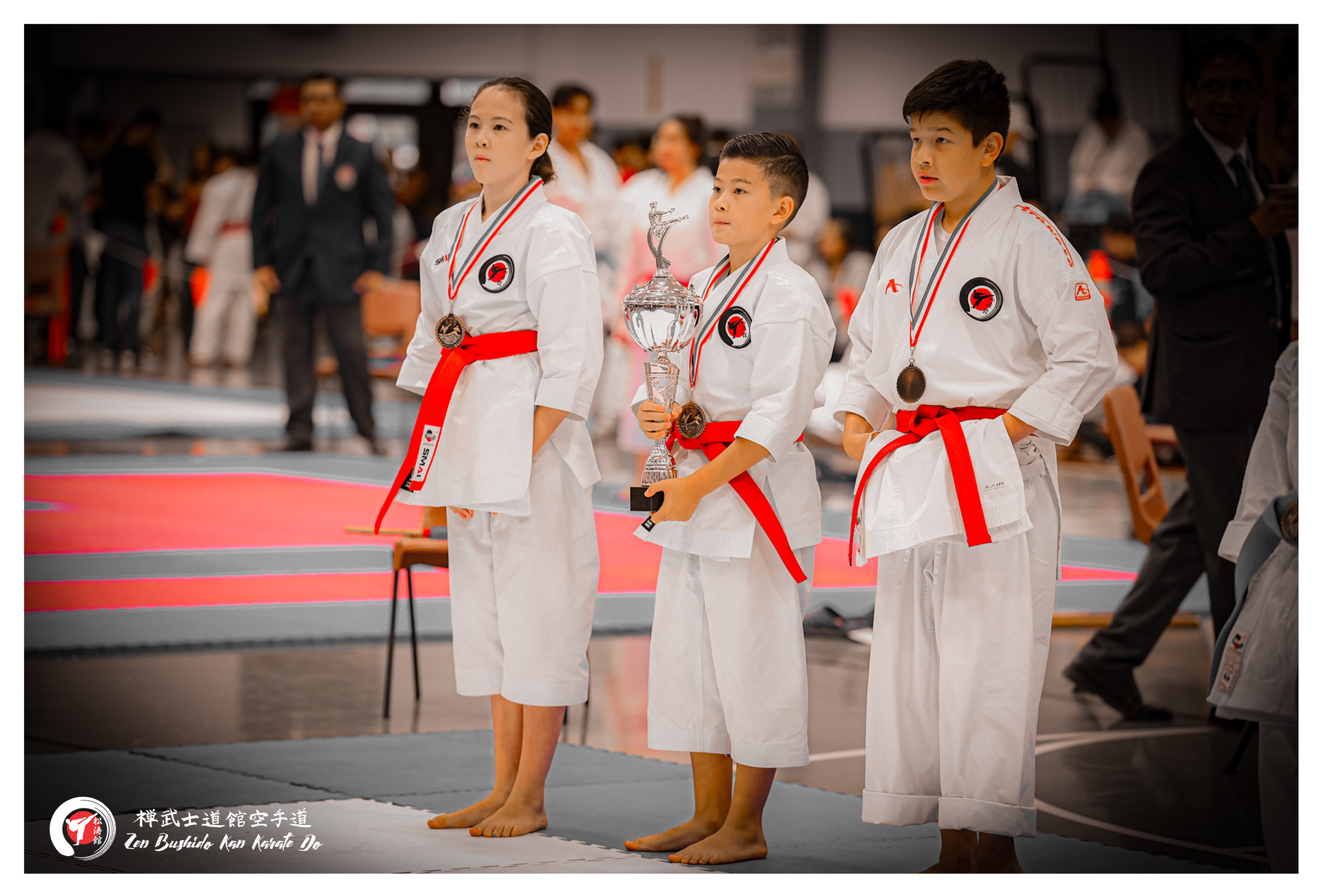 Three young karate students on a podium, one holding a trophy, all wearing white uniforms and red belts.