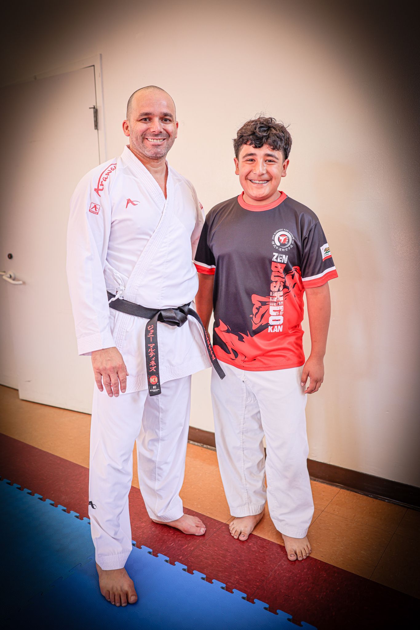 Two people in martial arts uniforms standing together indoors, smiling at the camera.