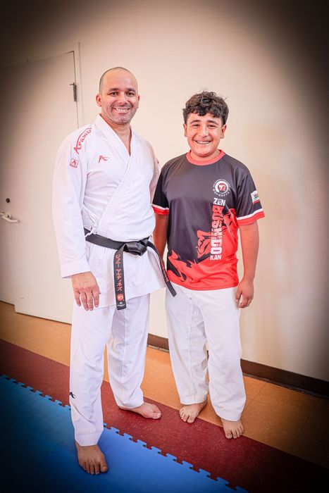 Two people in martial arts uniforms standing together indoors, smiling at the camera.