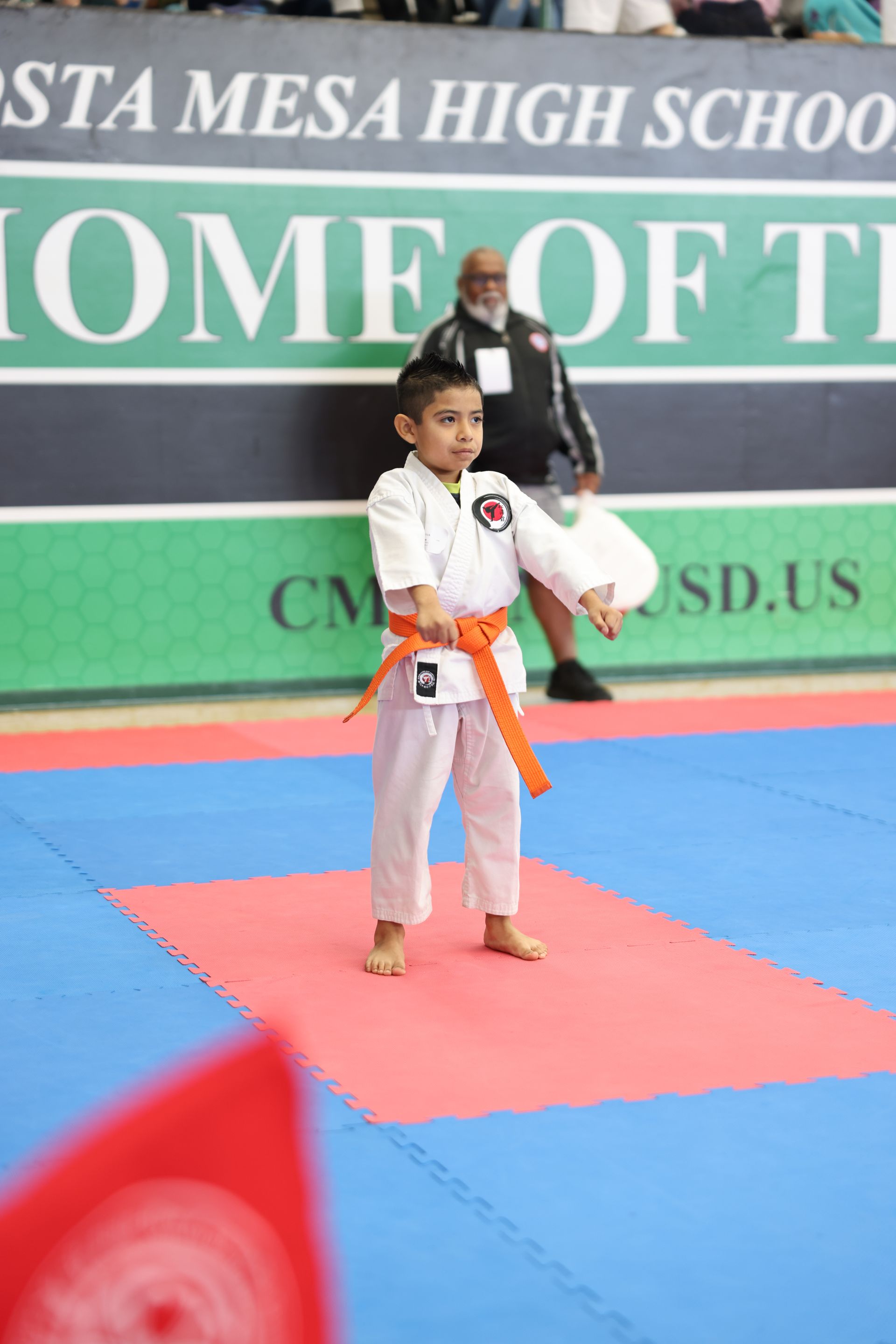 Young karateka in orange belt, performing a stance on a blue and red mat at a tournament.