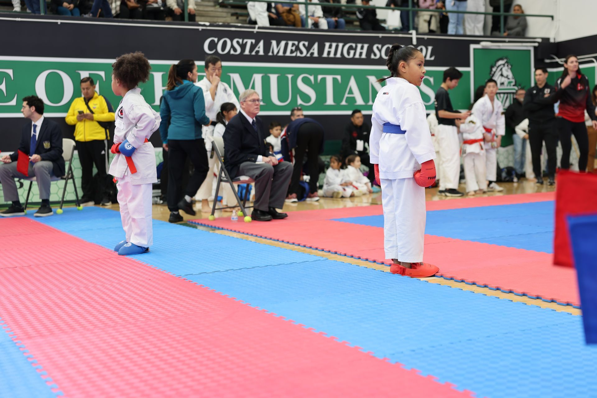 Two young karate competitors in white uniforms stand on a blue/red mat at Costa Mesa High School.