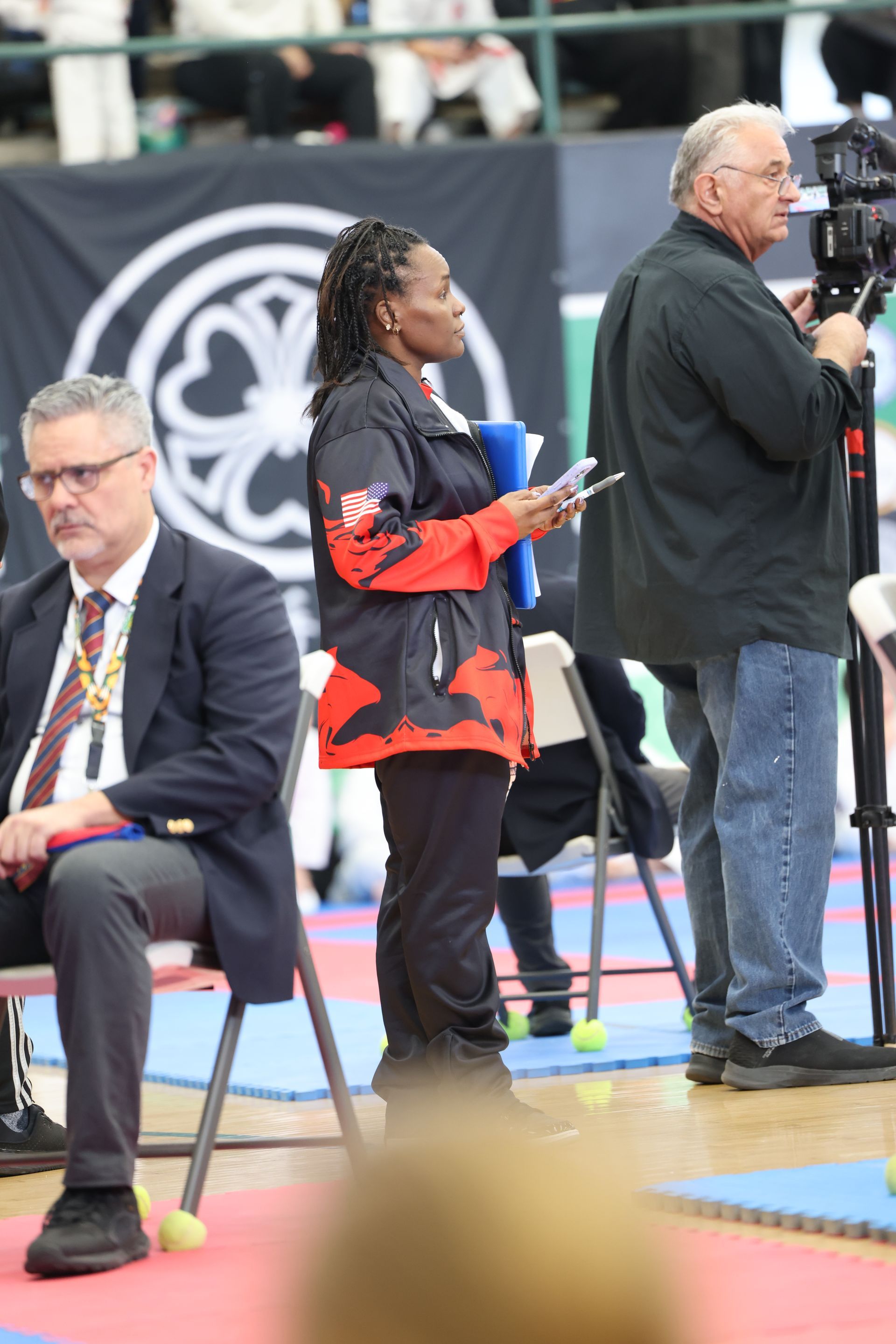 Woman in red/black jacket holding papers, stands near a man filming. Another man sits nearby. Competition setting.