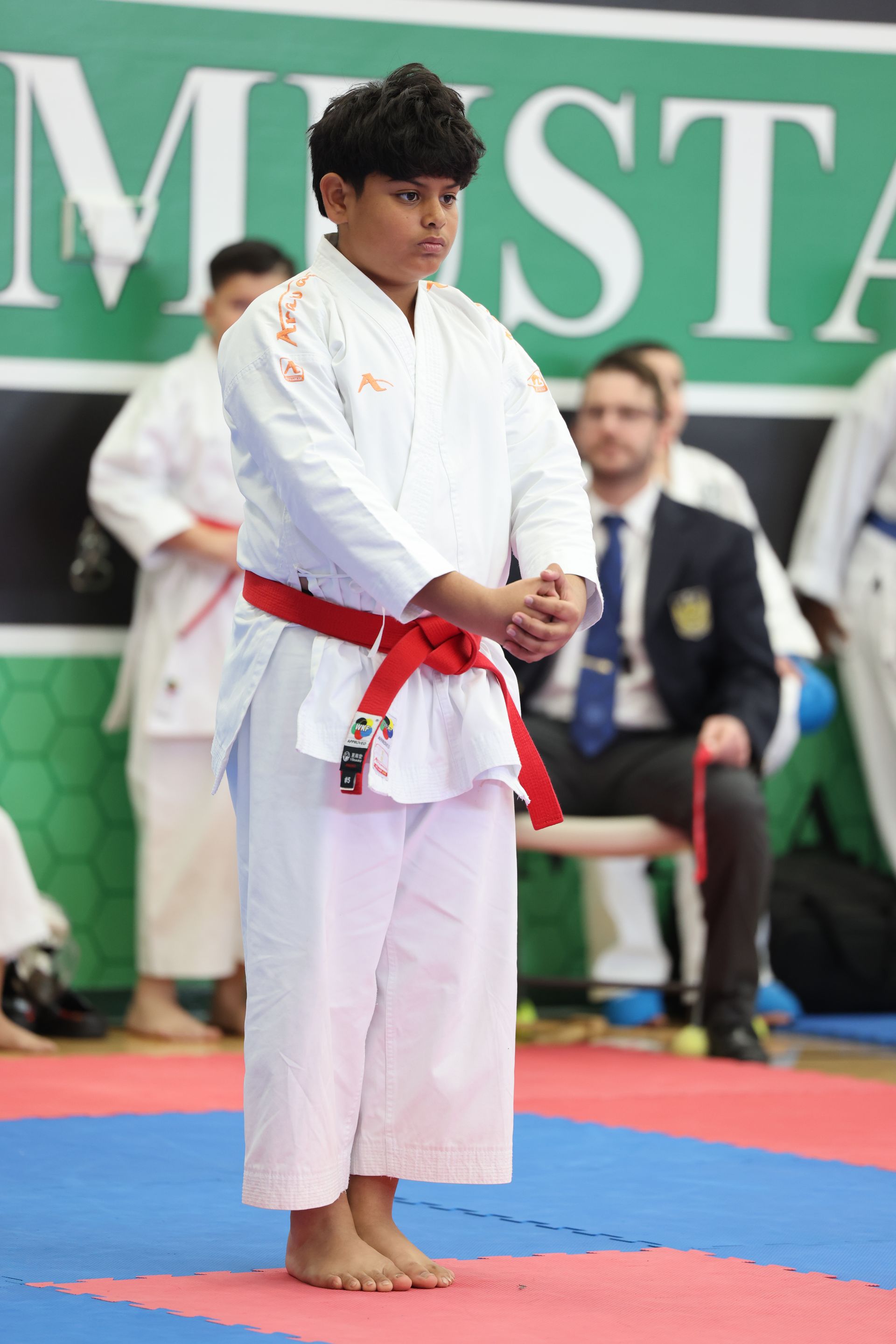 Karate practitioner in a white uniform with a red belt, standing on a blue and red mat.