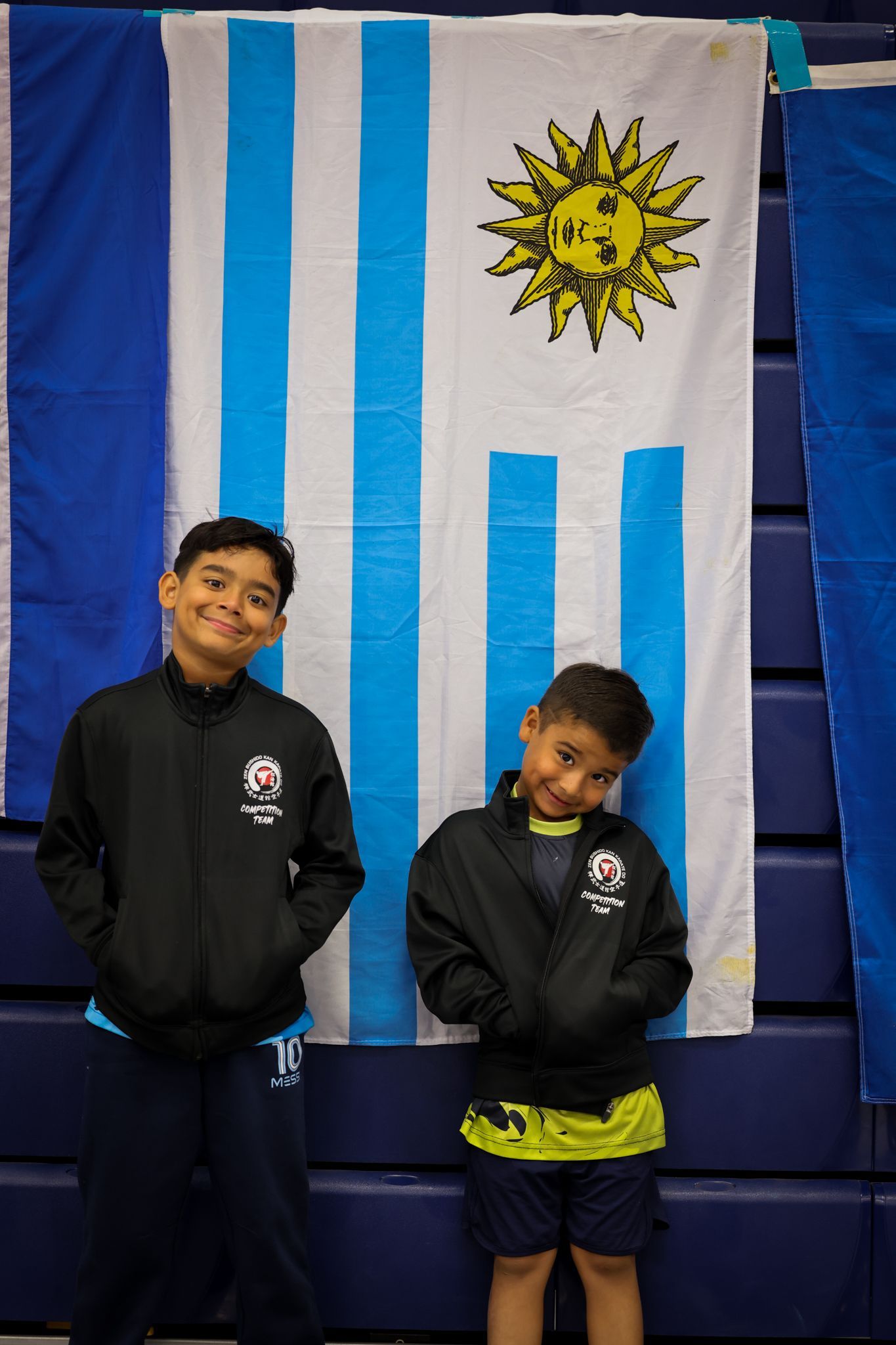 Two boys smiling in front of the Uruguayan flag, wearing black jackets and dark blue pants.