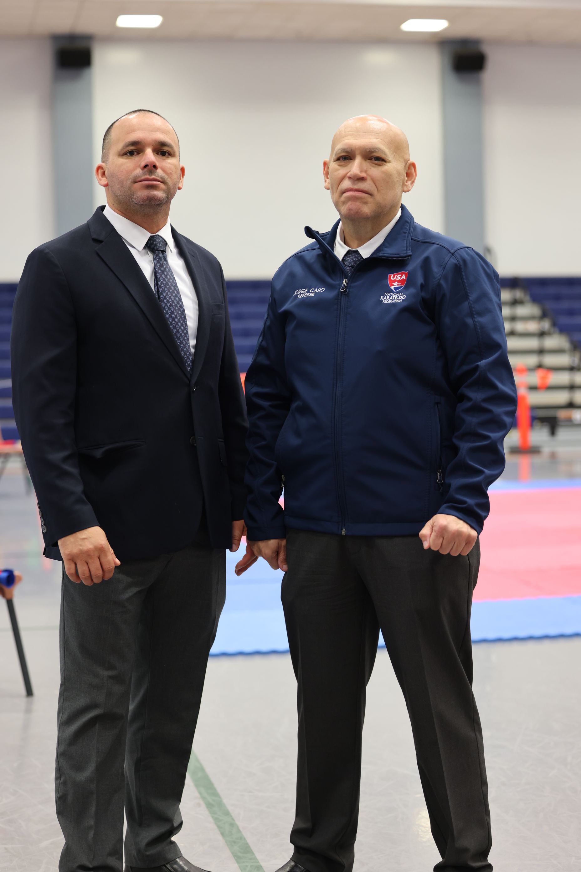 Two men, one in suit and tie, one in blue jacket, stand in a gymnasium, likely officials.