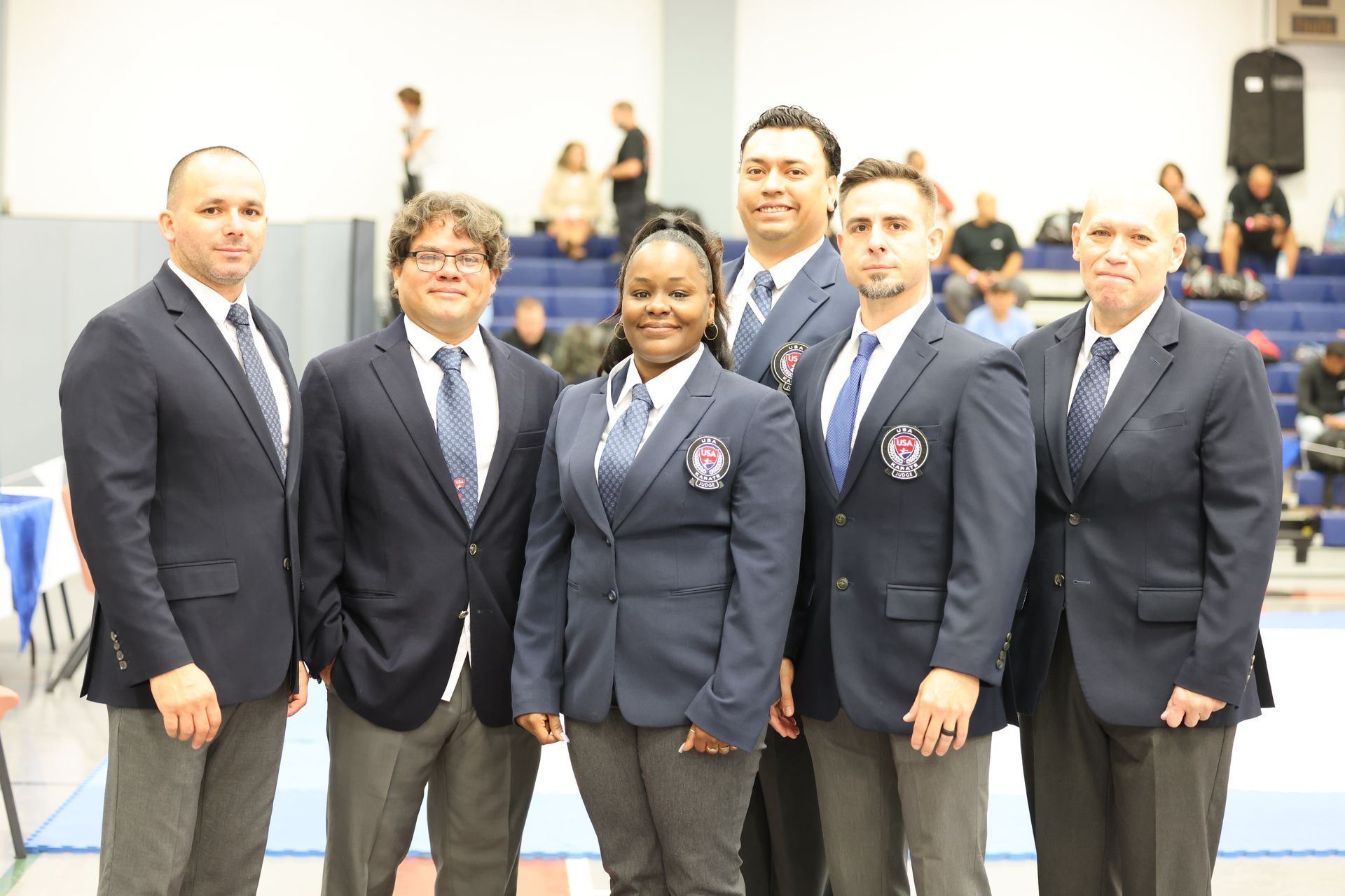Group of six referees in navy blazers and ties pose for a photo, indoors in a sports venue.