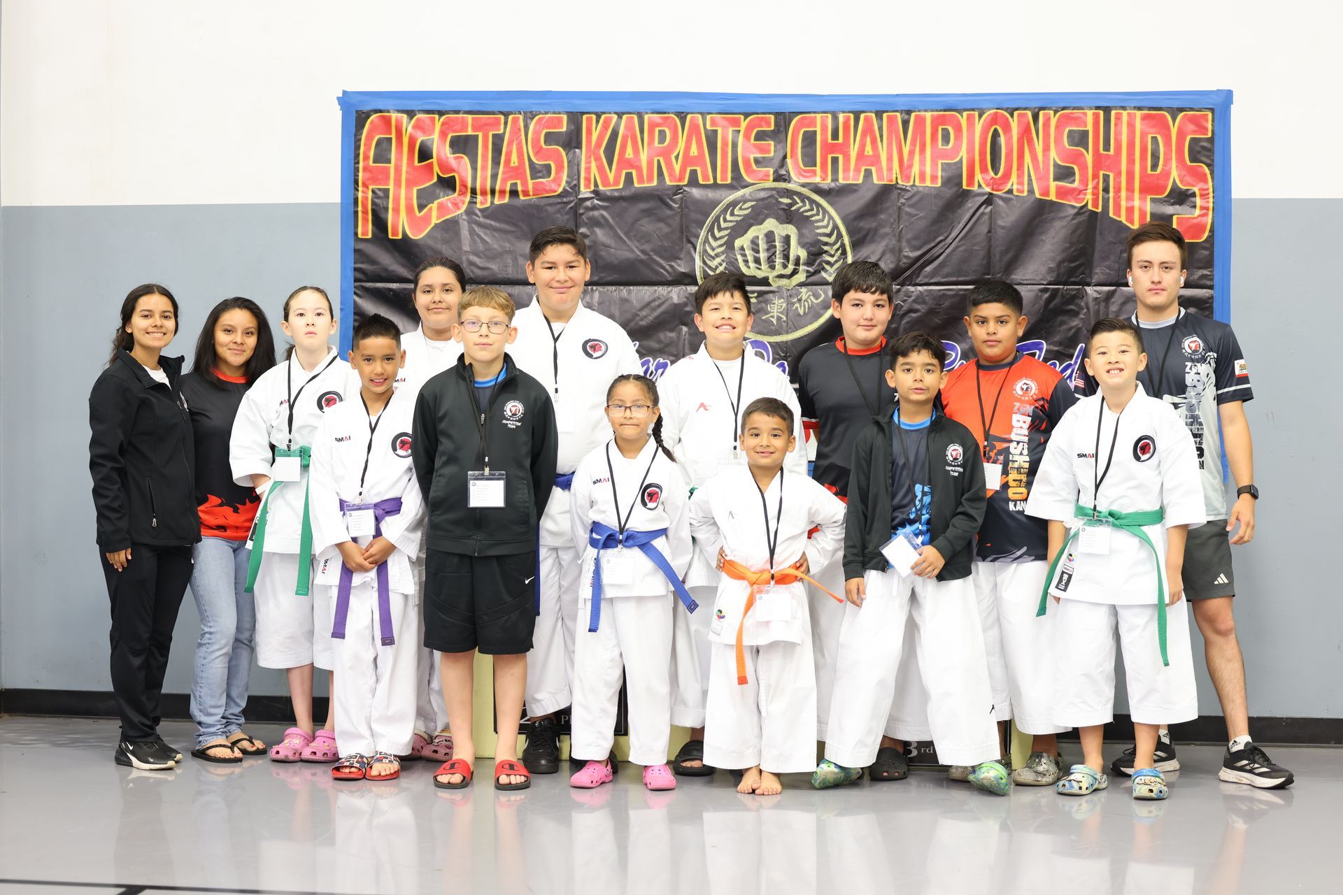 Group of young karate students posing with coaches at a tournament, banner in background.