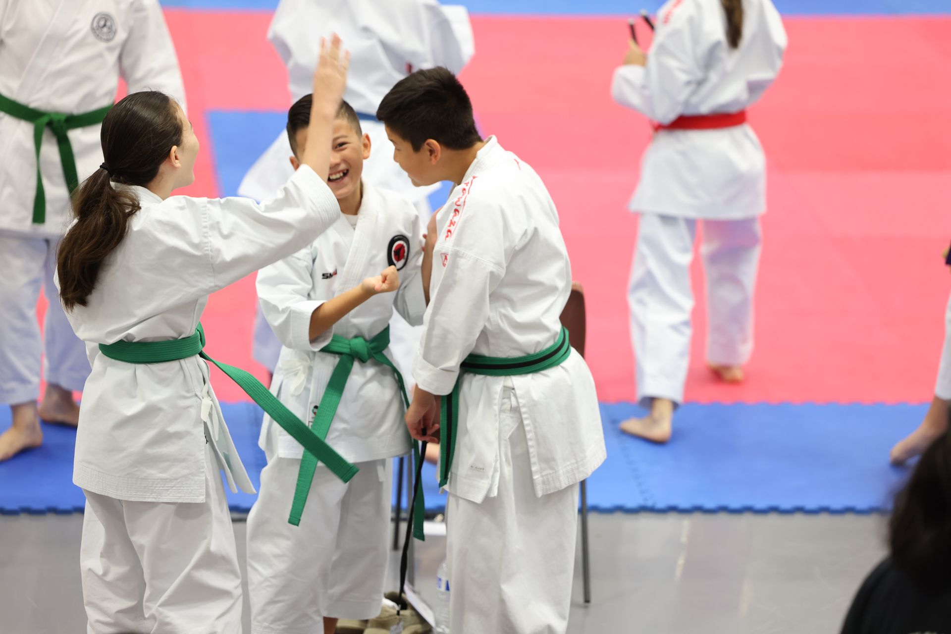 Three children in karate uniforms celebrate with high-fives at a competition.