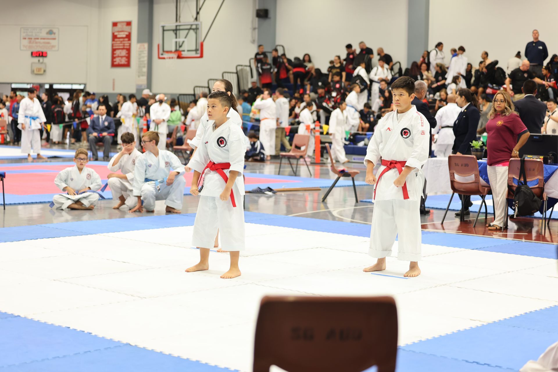 Two young karatekas in white uniforms with red belts face each other on a mat, ready to compete.