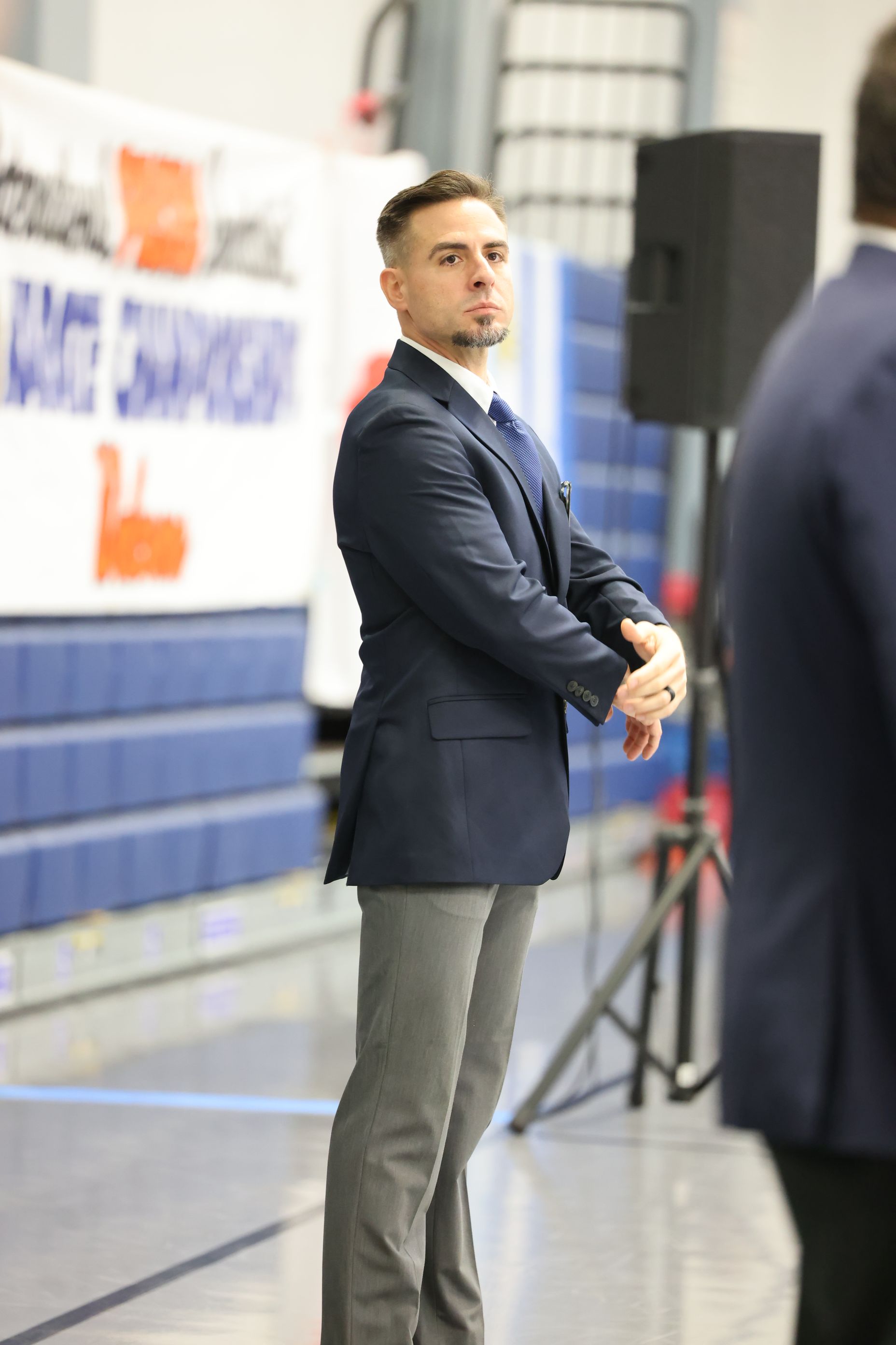 Man in a navy blazer and gray pants stands near a blue wall, looking to the side.