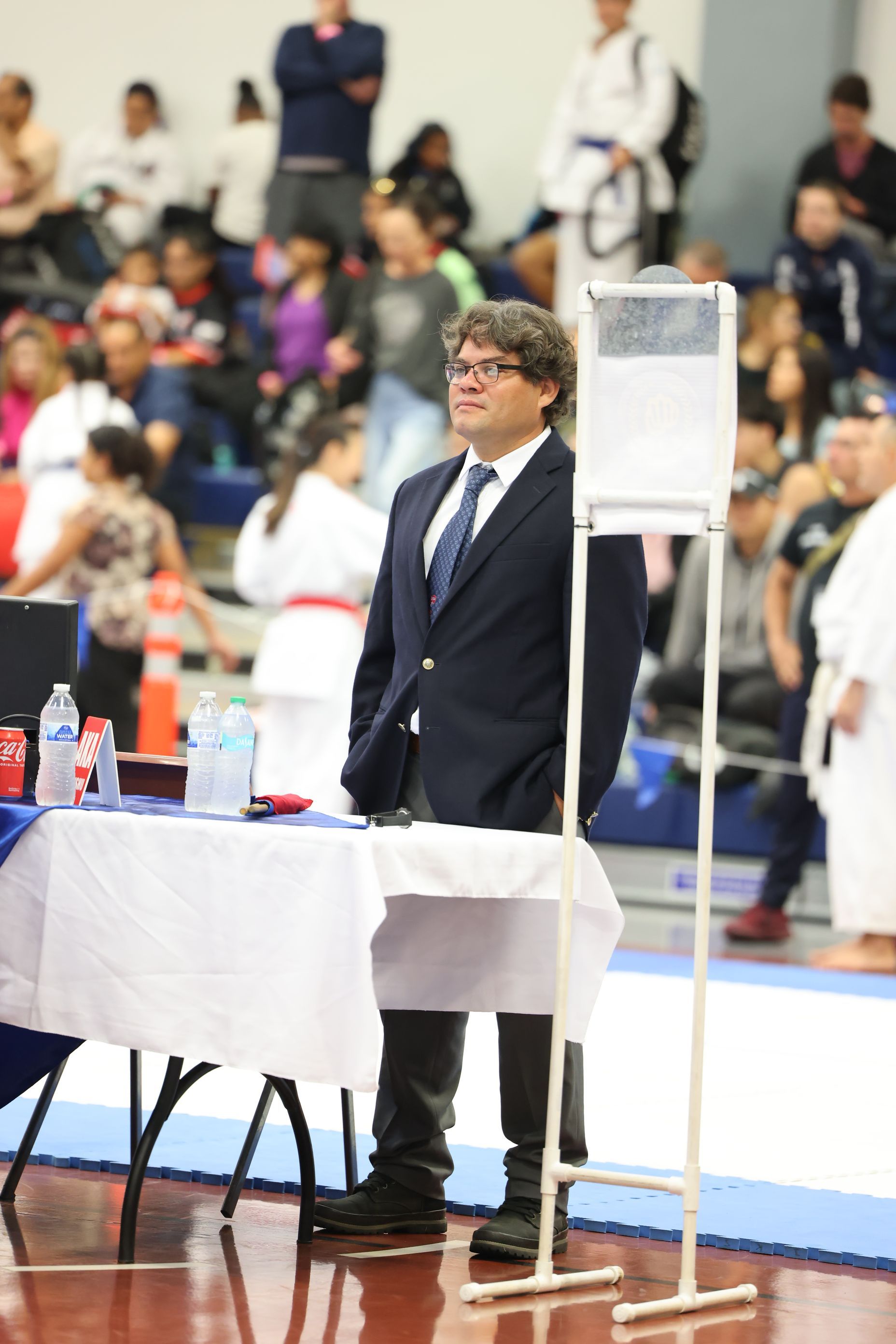 Man in suit at a sports event. He is standing by a table with a score display, observing the competition.