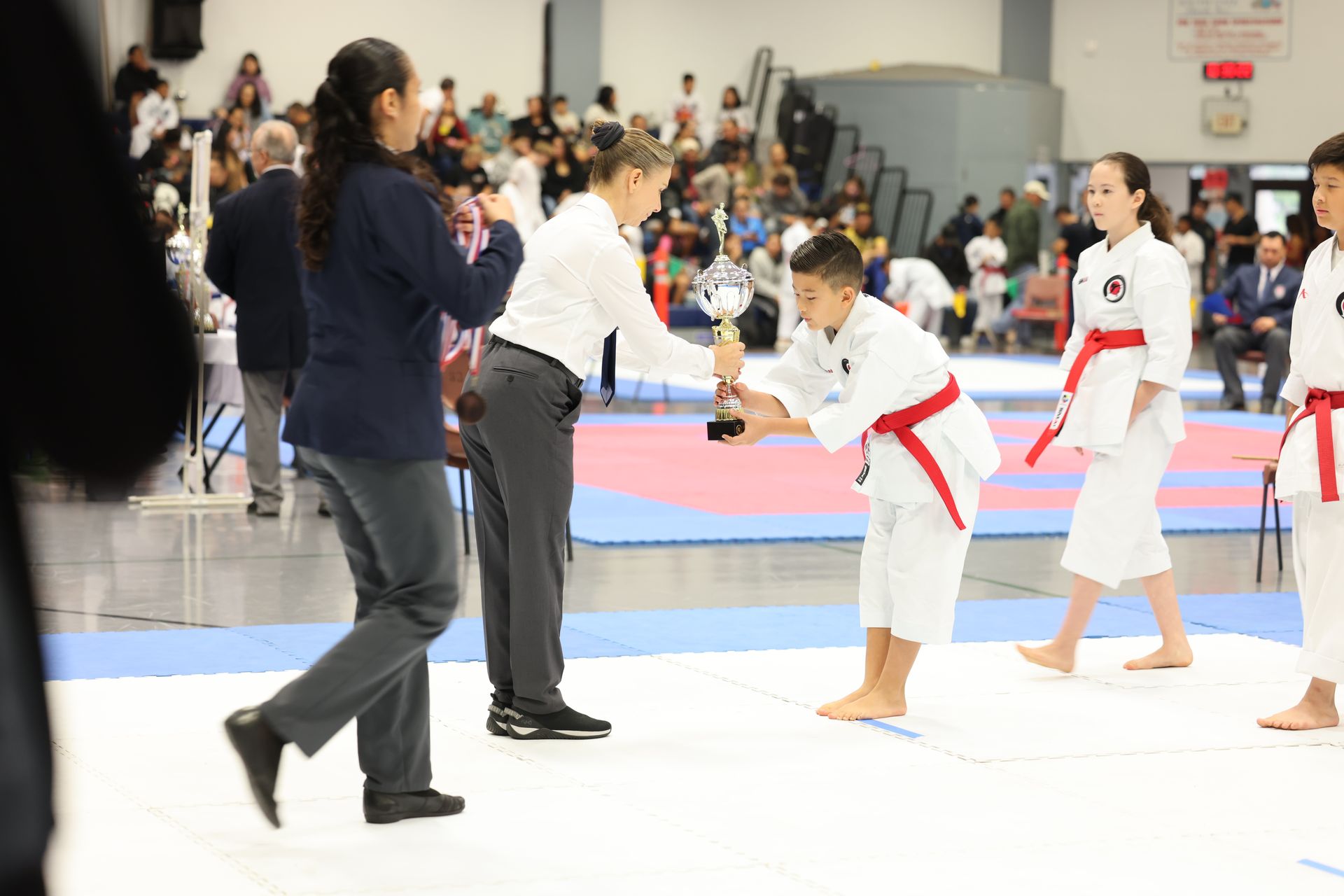 Boy in karate uniform receives trophy at a competition. Other competitors and judges in background.