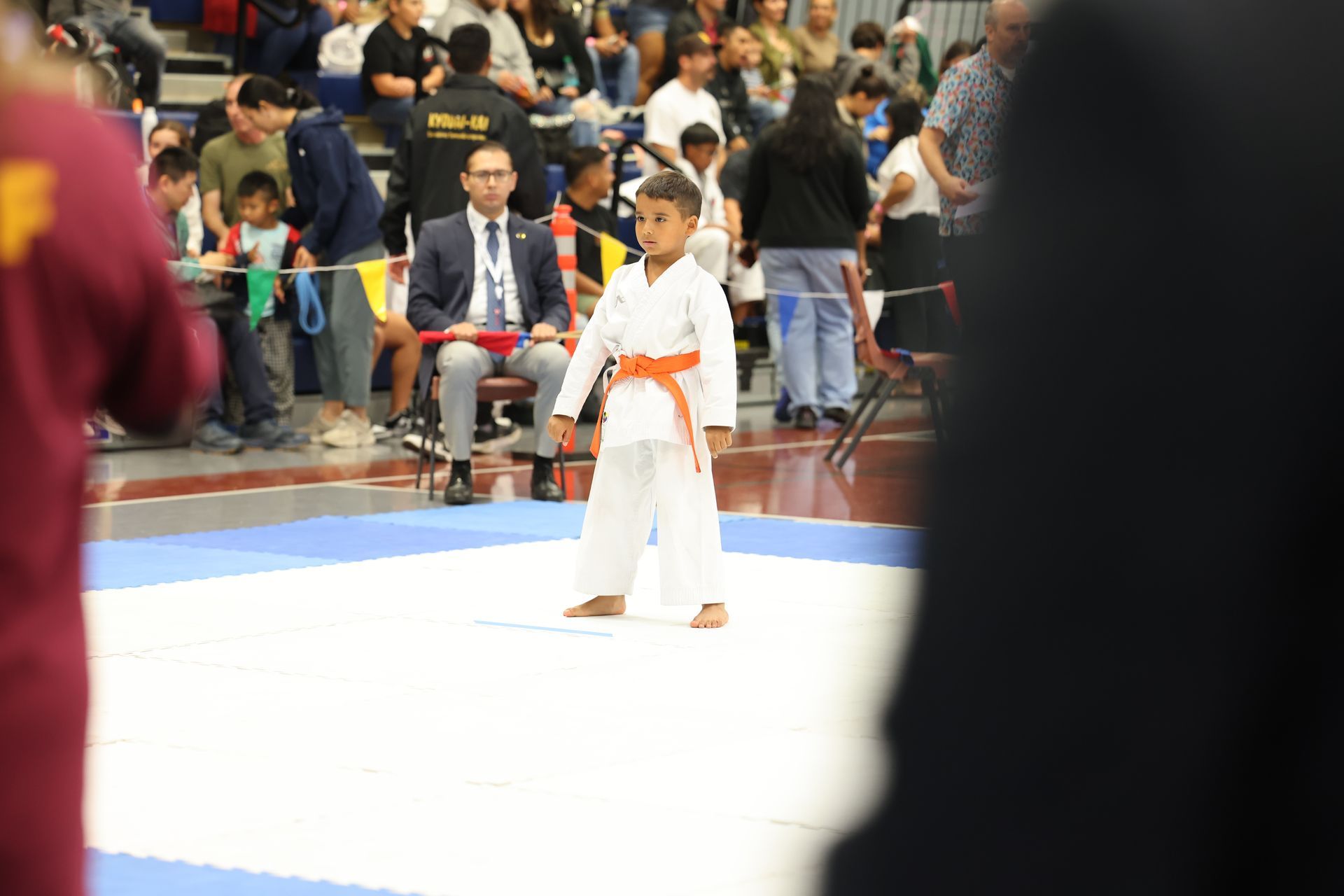 Young boy in karate uniform with orange belt stands on a mat at a tournament.