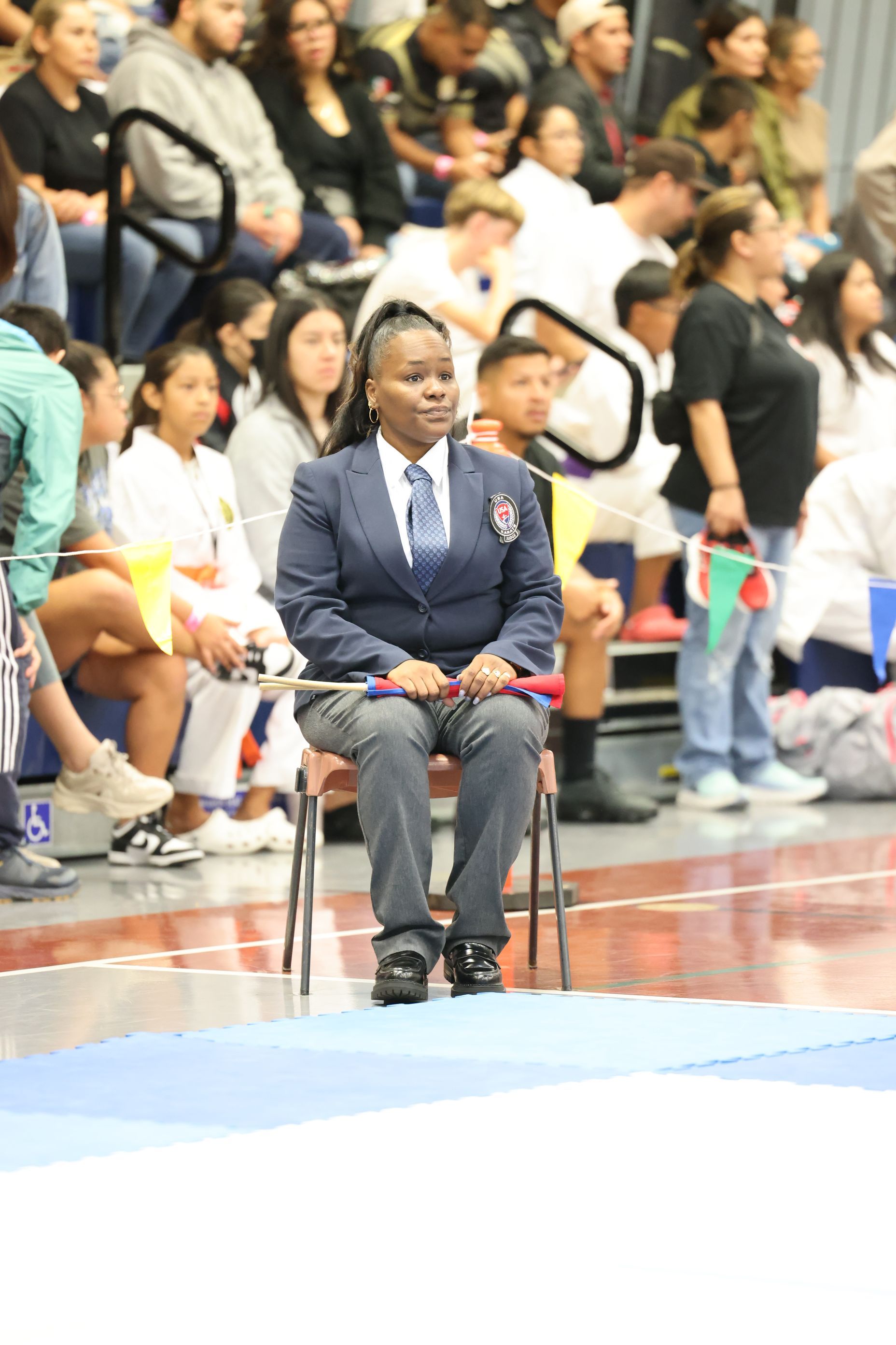 Woman in blazer and tie, seated, observing karate competition. Indoor gymnasium setting.