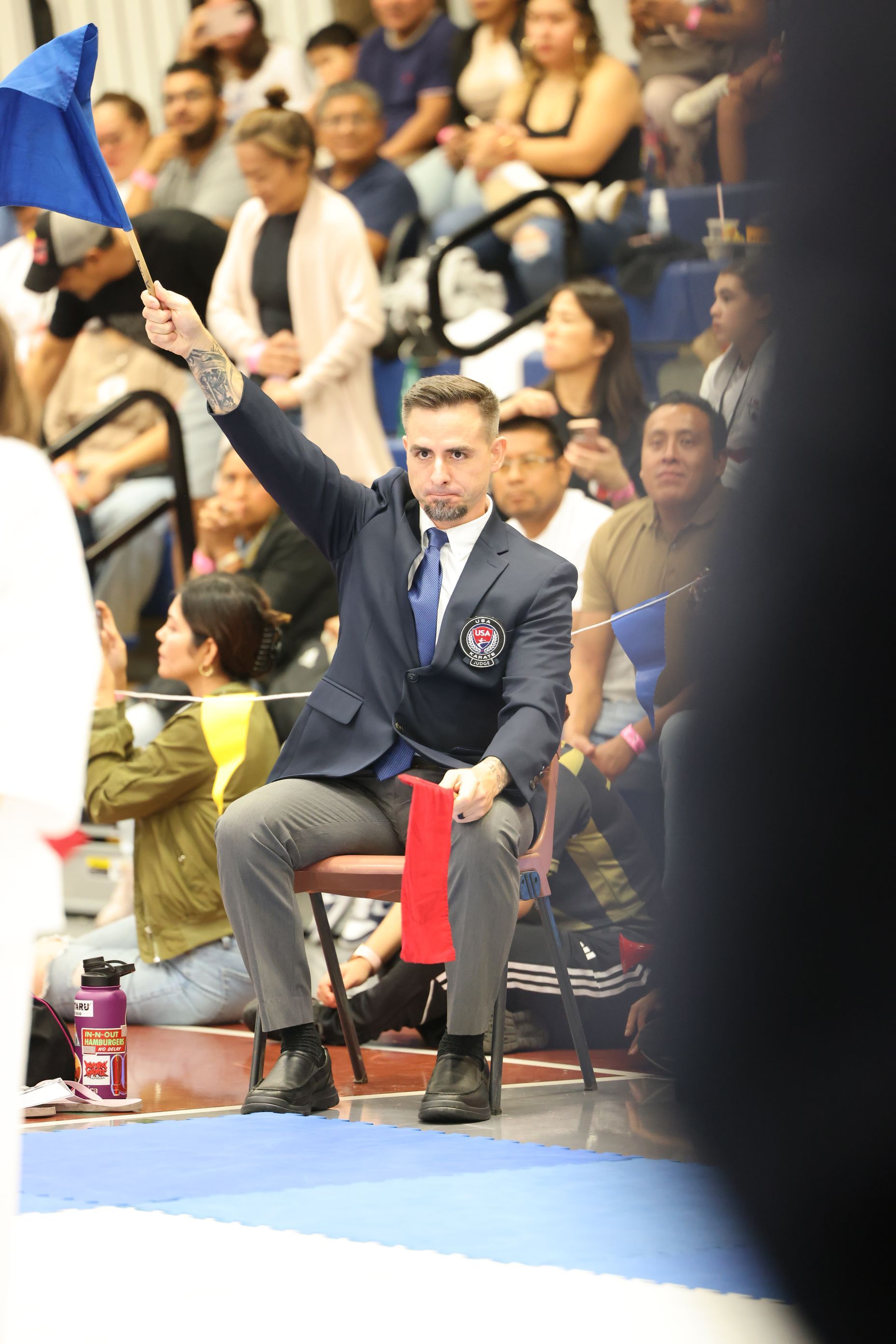 Referee in a blue blazer raises a blue flag at a sports event. People watch from bleachers in the background.