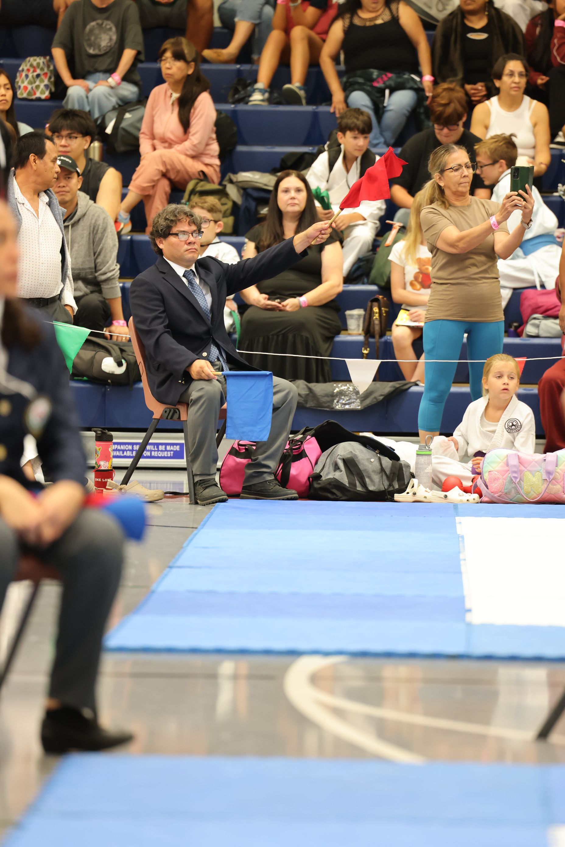 A man in a blazer coaches during a sports competition, gesturing toward the field. Spectators watch from the bleachers.
