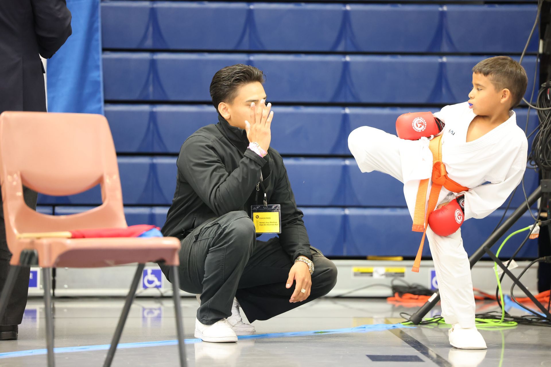Young karate student in white uniform kicks, watched by coach, indoor gym setting.