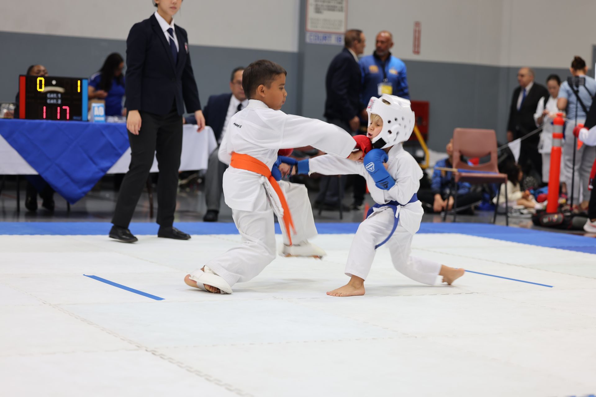 Two young karate athletes sparring; orange vs. blue. The referee watches the action in an indoor competition.