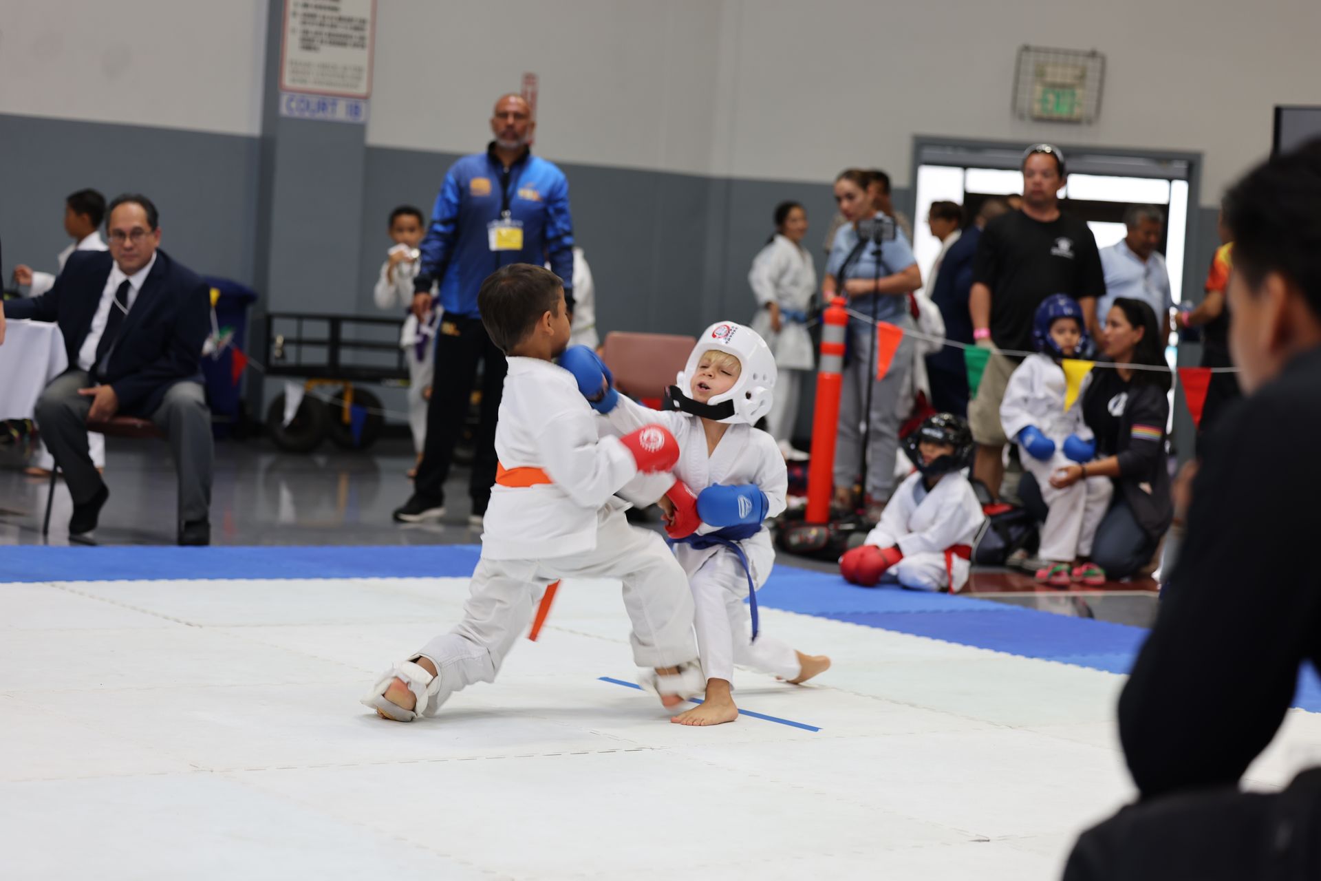 Two young boys in karate gear sparring at a tournament. One with an orange belt throws a punch. Others watch.